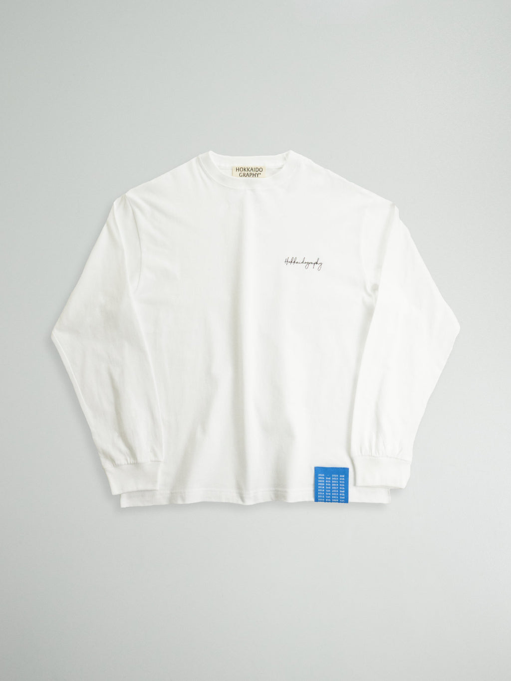 HOKKAIDOGRAPHYの新作「THE RECORD」ロンT（ホワイト）。左胸にロゴ、左裾に北海道のプロ野球の戦績を記した青いタグを配した全体像。