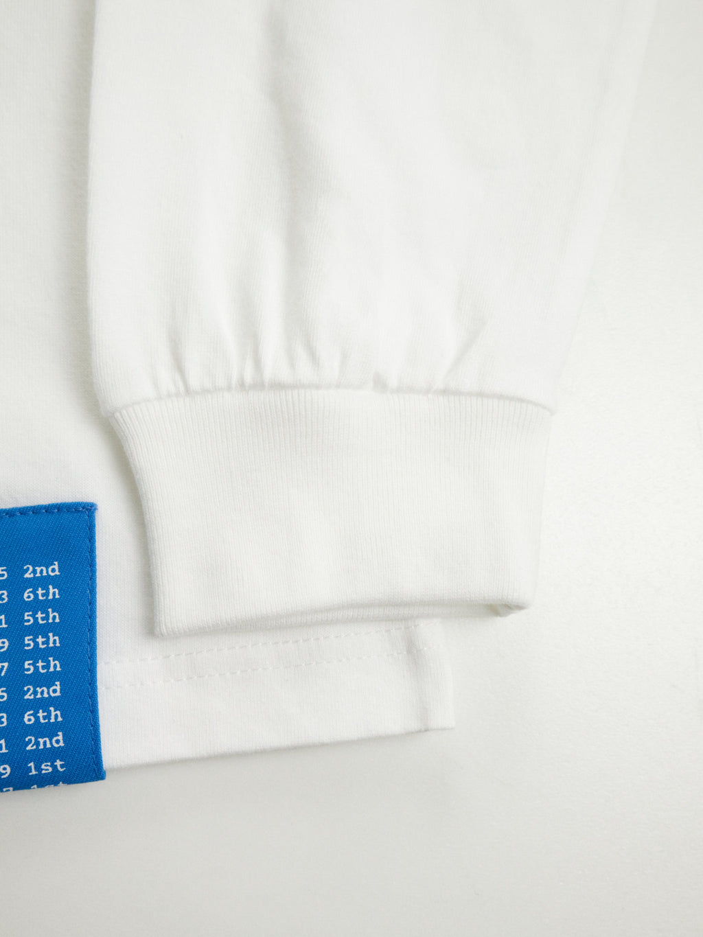 6.2オンスのタフな生地を使用したロングスリーブTシャツの袖リブと、左裾の青い戦績タグの接写。